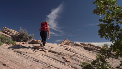 Espalda-De-Mujer-Con-Mochila-Caminando-Sobre-Una-Colina-Rocosa-En-El-Desierto-Bajo-El-Cielo-Azul,-Cámara-Lenta