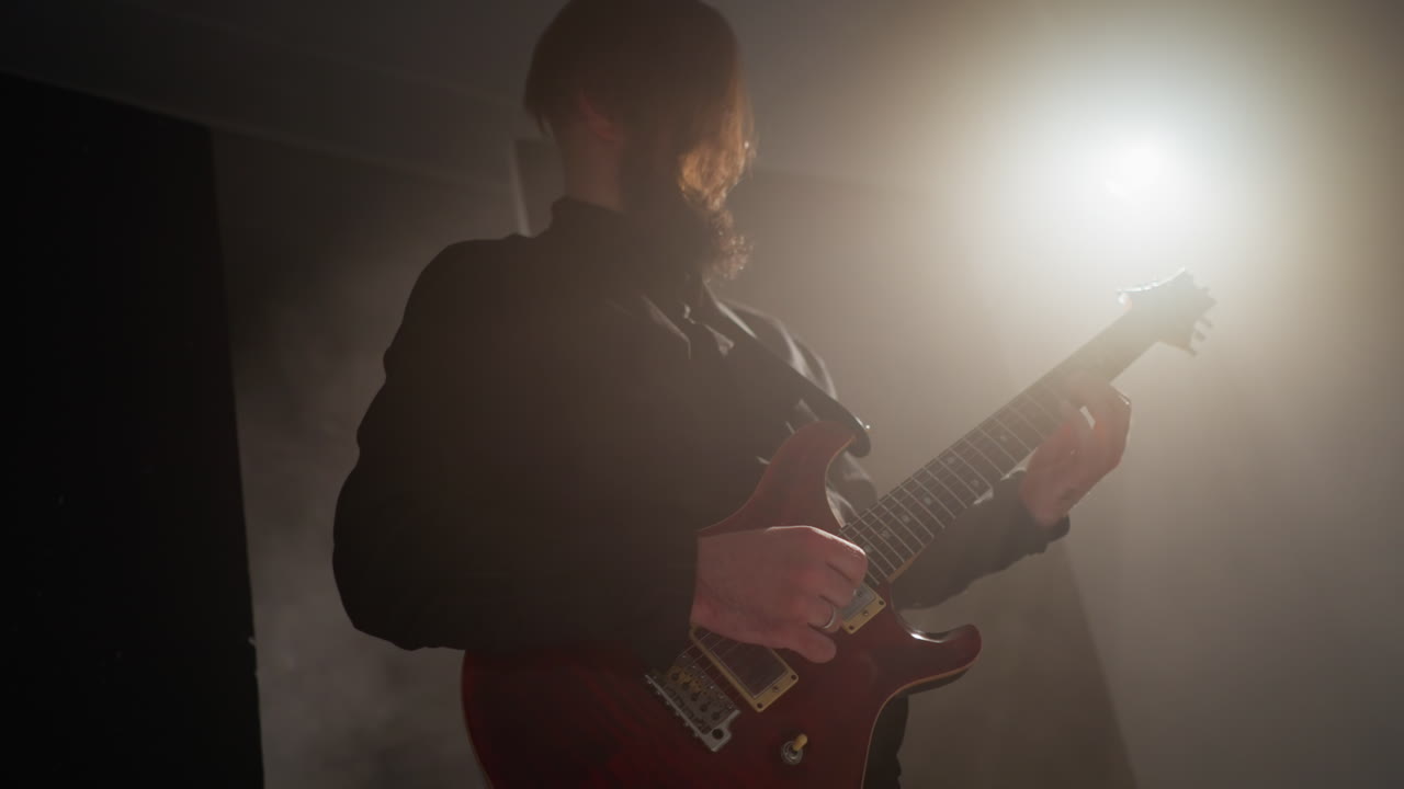Guitarist in a black outfit playing a red guitar with expressive emotion, lit by a single spotlight in a dark room