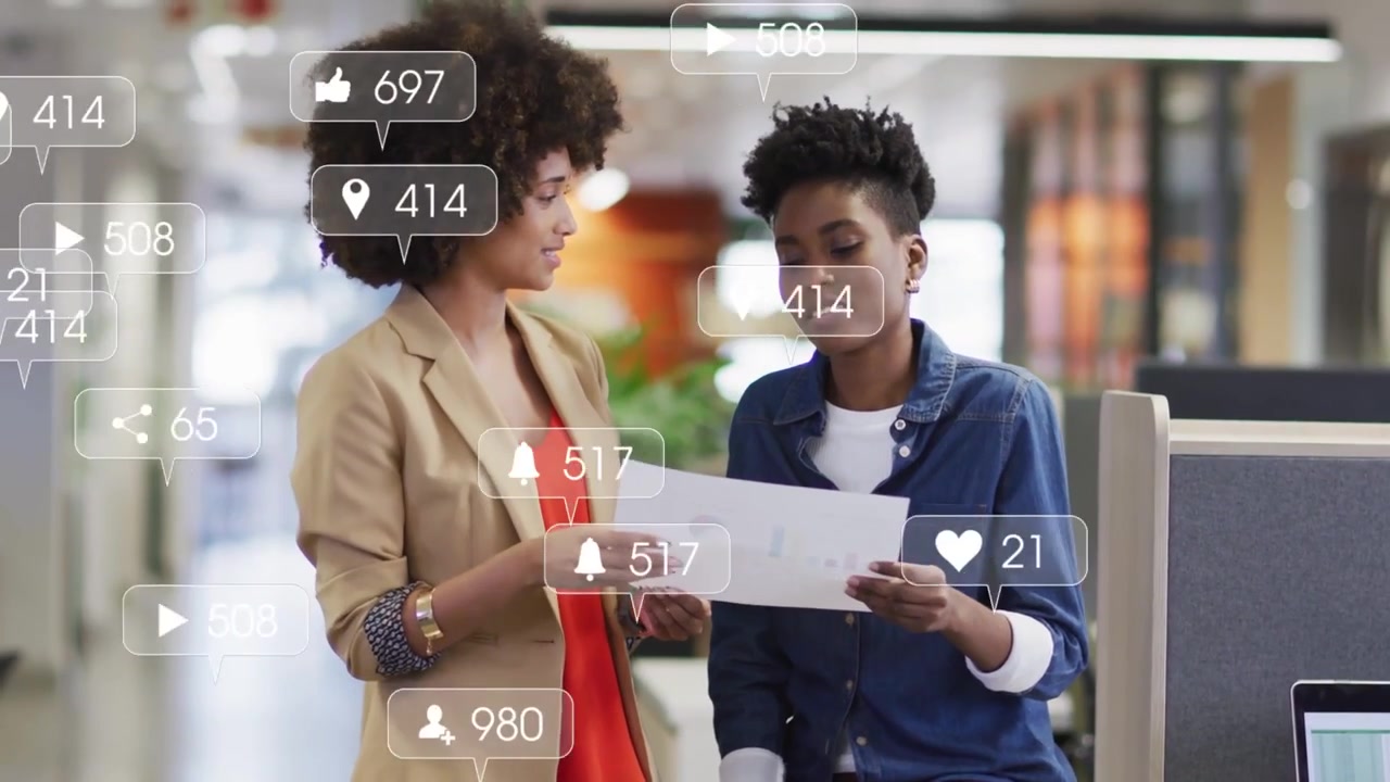 Two women handing paper at desk reviewing document for marketing as social alerts popping on laptop
