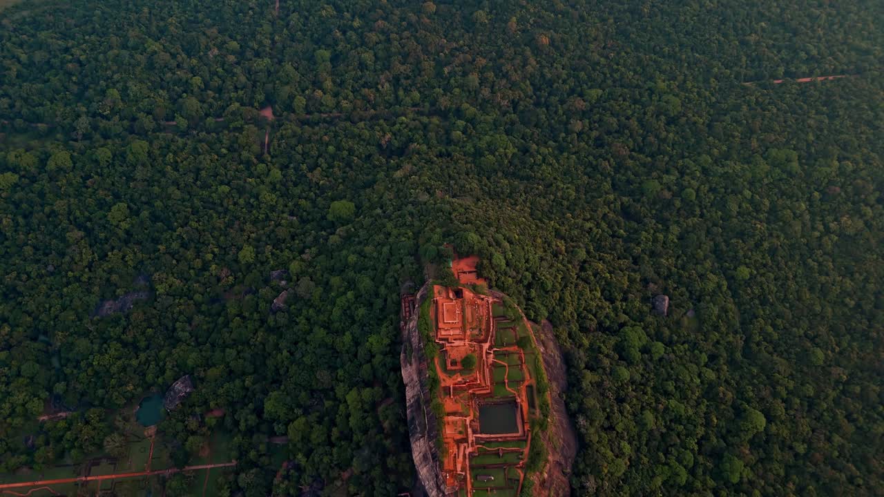 High-angle aerial drone footage directly above Sigiriya Rock Fortress at sunrise, capturing the symmetrical design of the ancient ruins, surrounded by lush jungle and glowing morning mist.