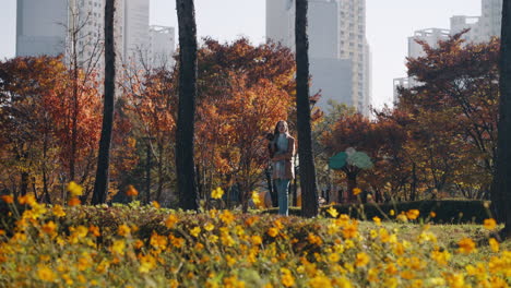 young blond woman holding toy poodle dog in her arms walking in autumn city park, yellow flowers in foreground - pan right slow motion