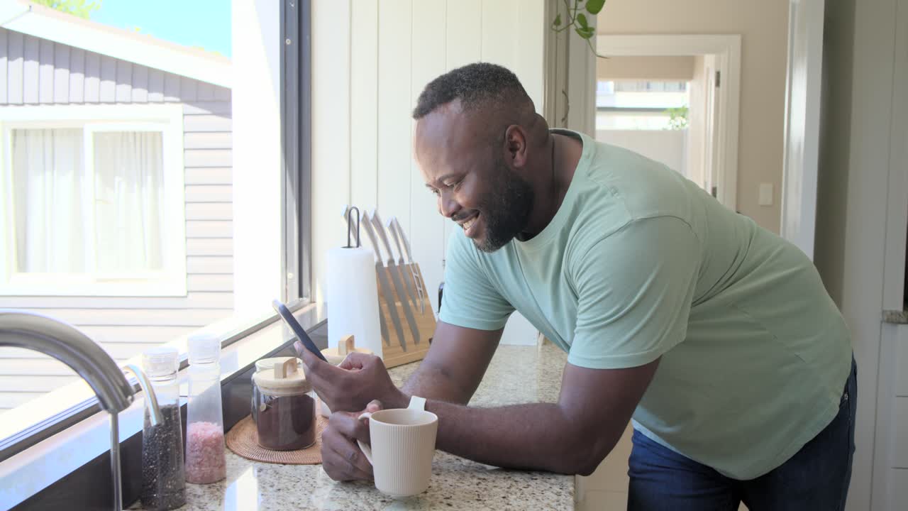 African American man receiving funny text, scrolling phone and sipping coffee on kitchen counter