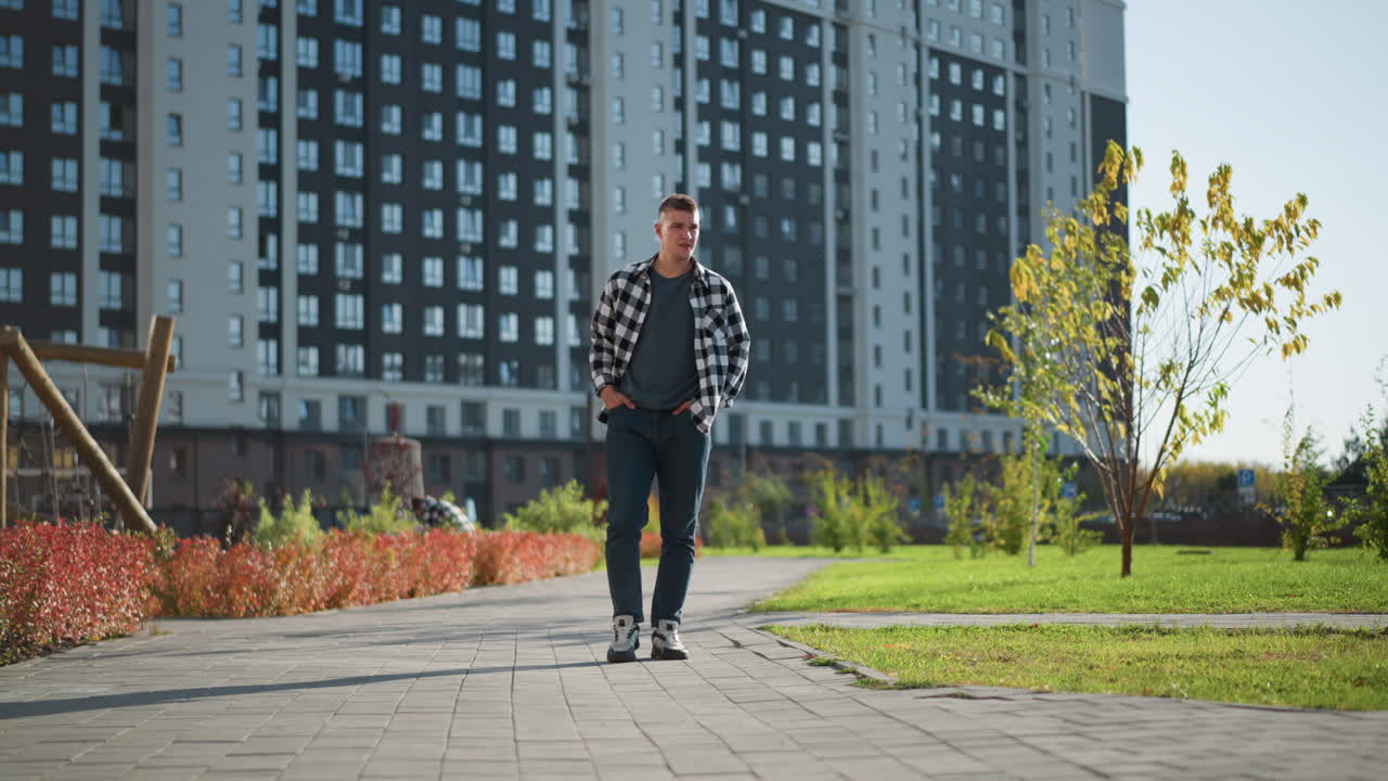 young man in black and white checkered shirt strolling with hands in pockets on paved walkway beside green lawn and red shrubs with modern residential buildings and clear sky