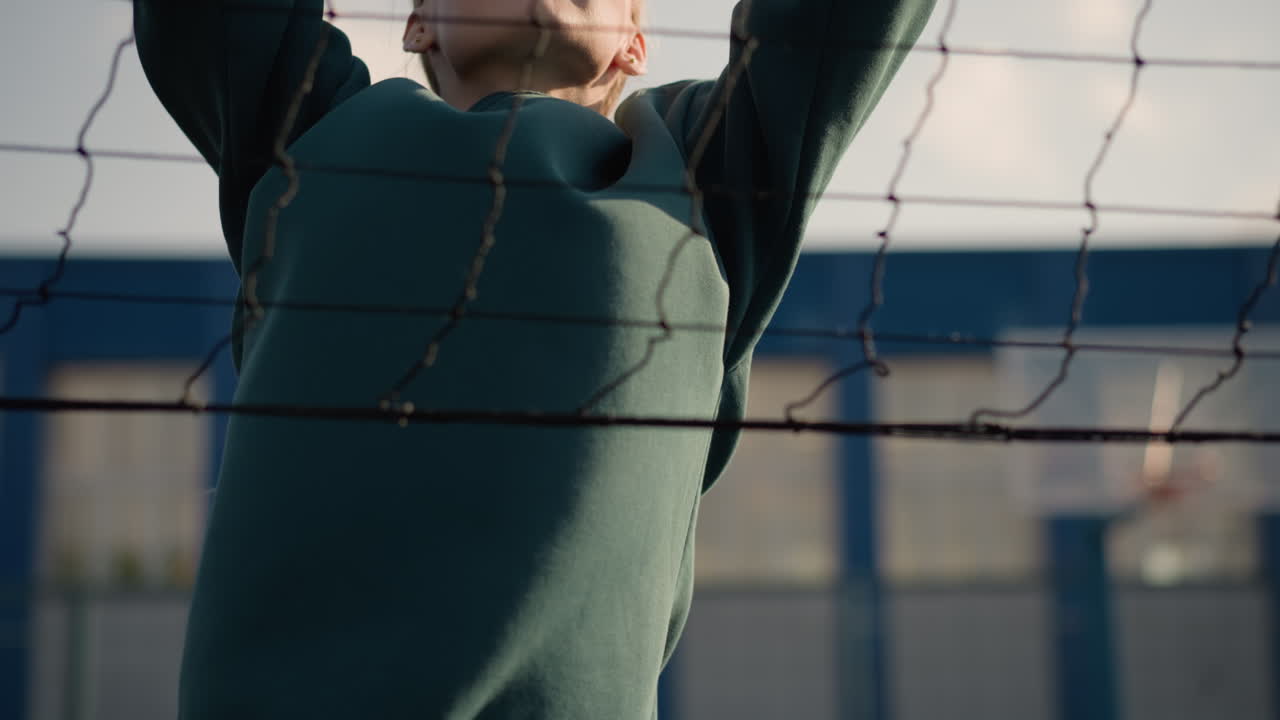 primer plano de una mujer con capucha verde haciendo ejercicio al aire libre, saltando con las manos levantadas cerca de la red, el fondo presenta un edificio borroso y un aro de baloncesto