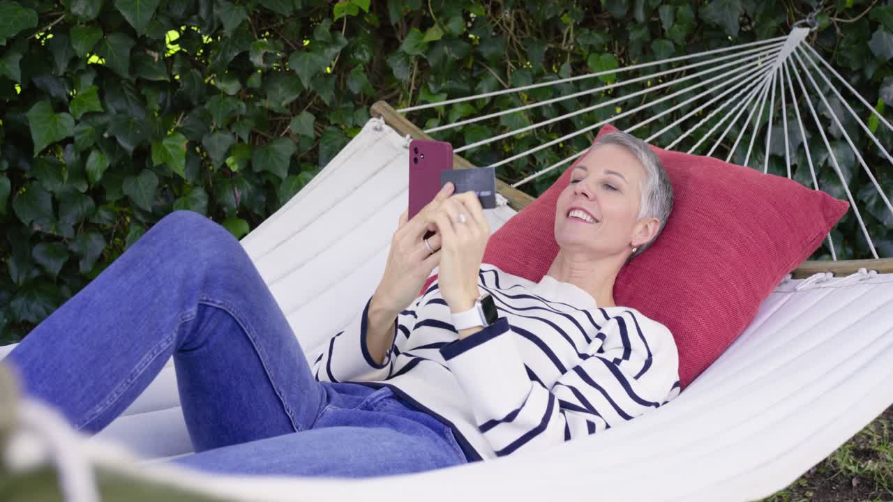 Sollecitata da un avviso di pagamento, una donna anziana solleva lo smartphone sull'amaca in giardino, toccando la carta per pagare.