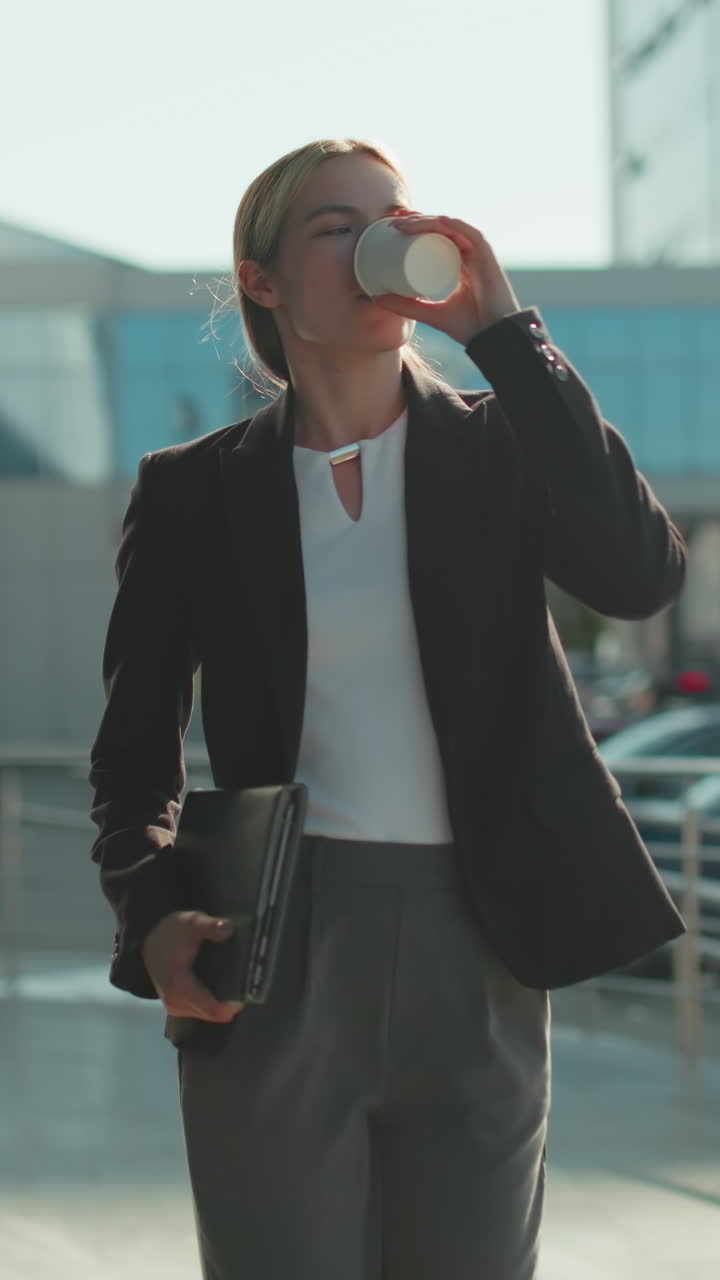 Elegant lady in black suit holding notepad in right hand and tea cup in left hand walks confidently with warm smile by modern glass building with parked cars and sunlight casting long shadow