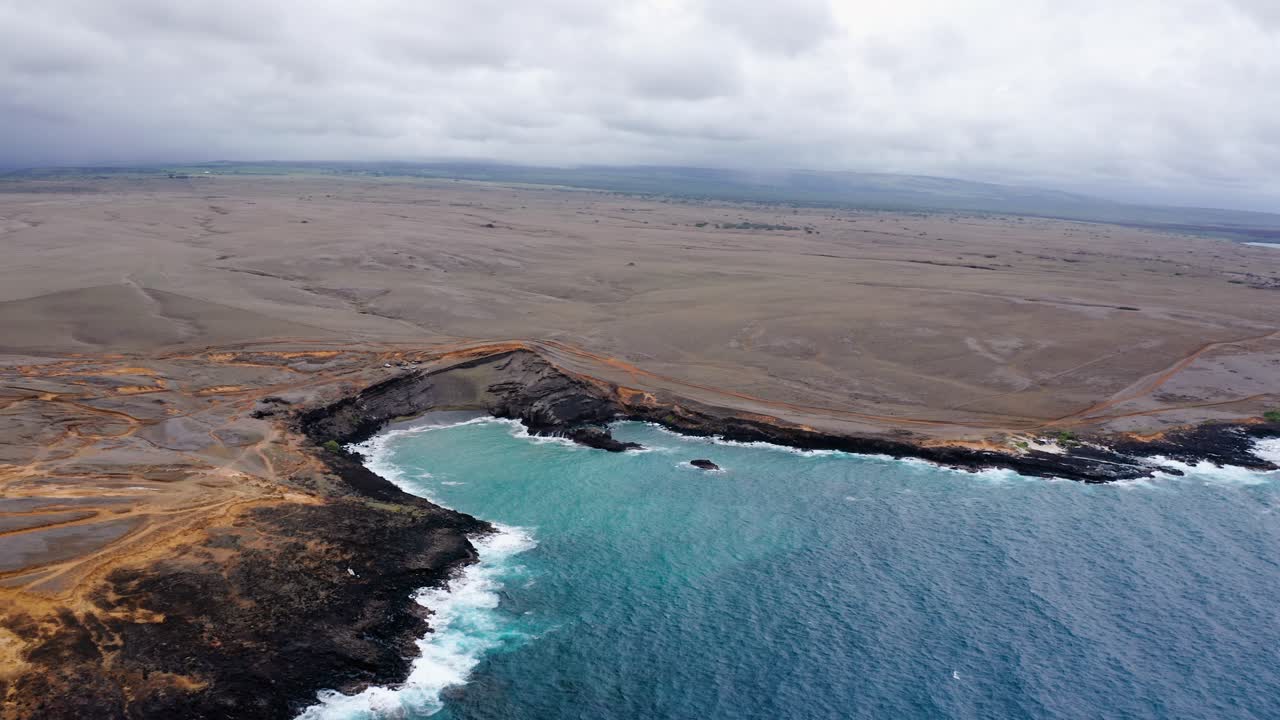 Aerial view of a rugged volcanic shoreline where vivid turquoise waves meet dark rocks. Vast barren land stretches inland under a moody, overcast sky.