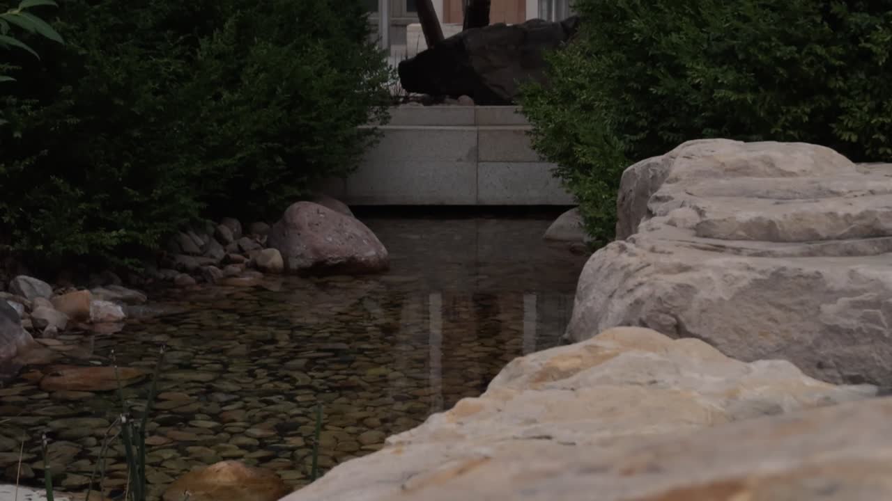 Low stream flows under stone walkway in landscaped garden with bushes and rocks.