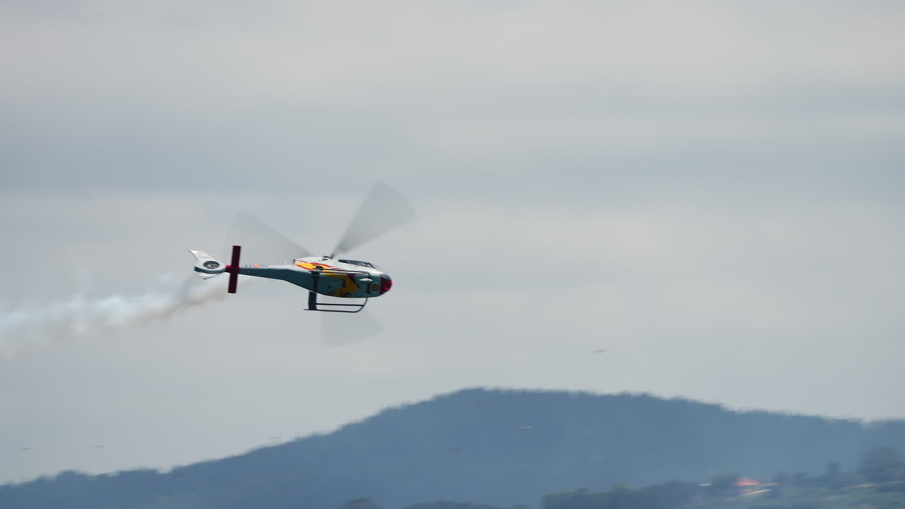Skilled helicopter pilot executing precise aerobatic maneuvers, generating dramatic white smoke trail against expansive sky and distant landscape