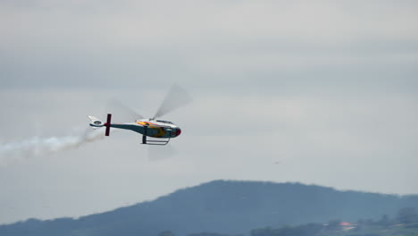 skilled helicopter pilot executing precise aerobatic maneuvers, generating dramatic white smoke trail against expansive sky and distant landscape