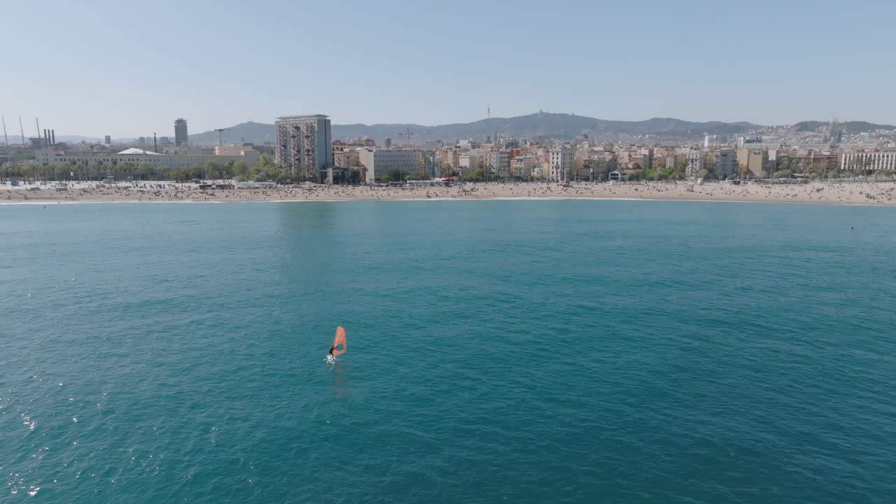 Slow drone approach from the Mediterranean towards San Sebastian and Barceloneta beaches in Barcelona, with a windsurfer gently sailing near the shore on a clear spring day