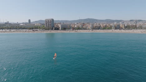 Slow drone approach from the Mediterranean towards San Sebastian and Barceloneta beaches in Barcelona, with a windsurfer gently sailing near the shore on a clear spring day