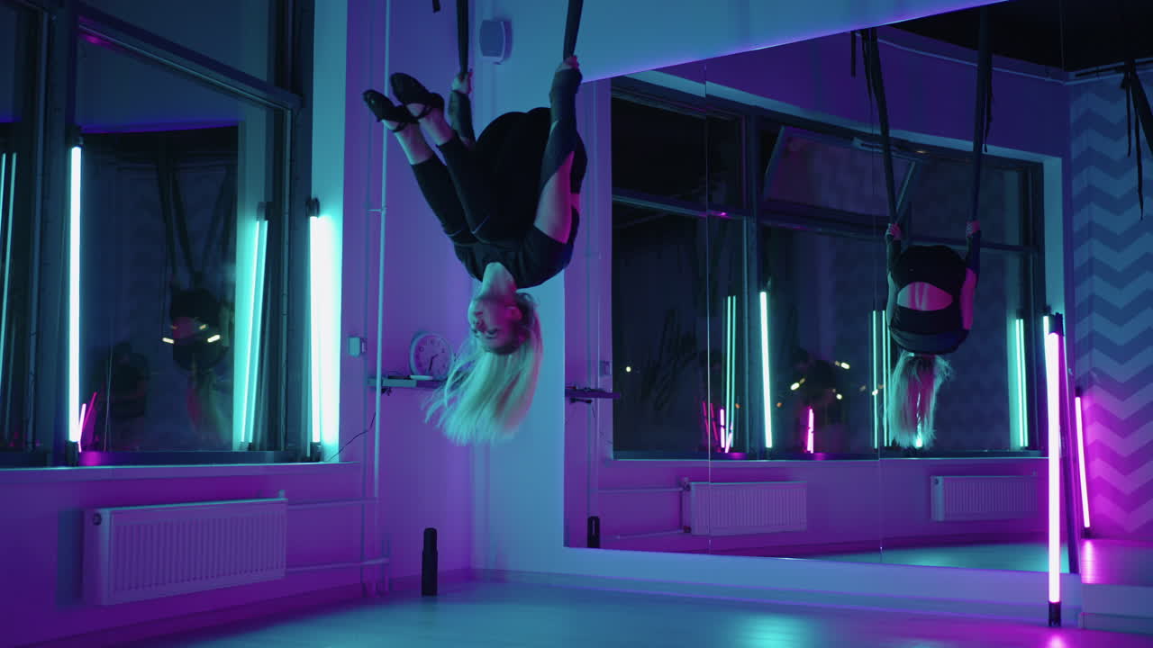 A woman does yoga and stretching on a hanging hammock in neon light. Young pretty slim body fitness girl practicing fly yoga in the gym.