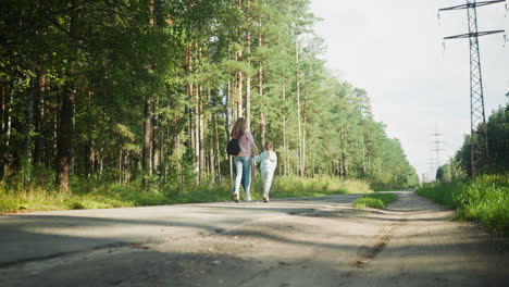 rear view of young lady joyfully holding hands with two siblings as they run along quiet cracked forest road surrounded by tall trees, green foliage, and morning sunlight in peaceful countryside