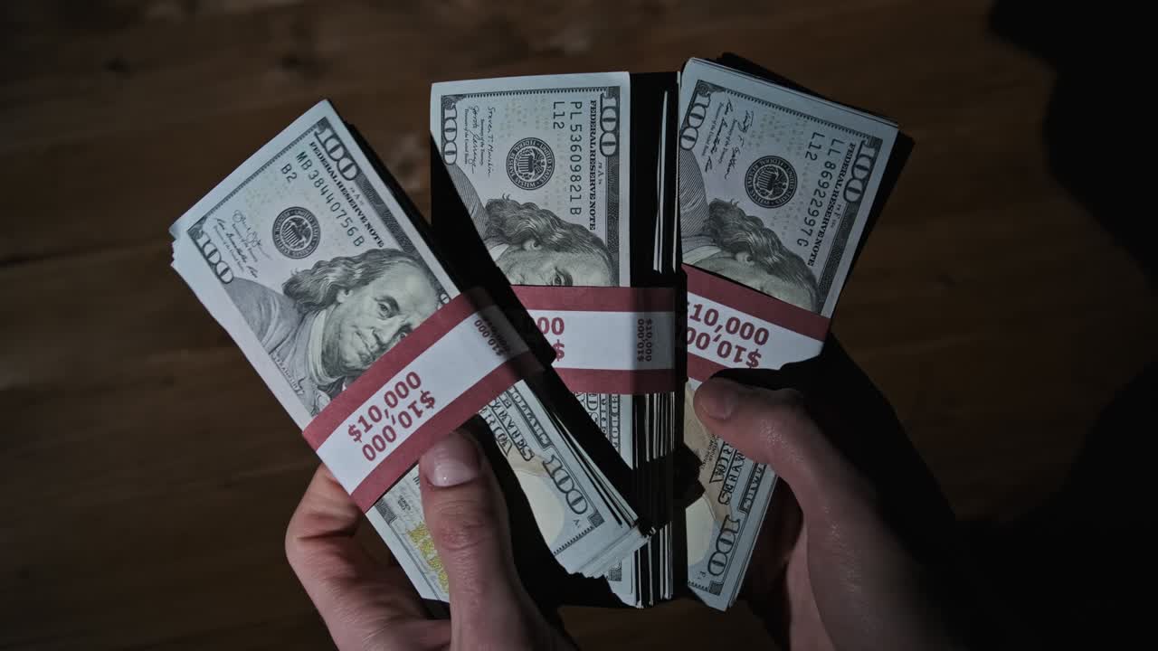 las manos masculinas sostienen tres pilas de billetes de 10.000 dólares en un fondo de madera.