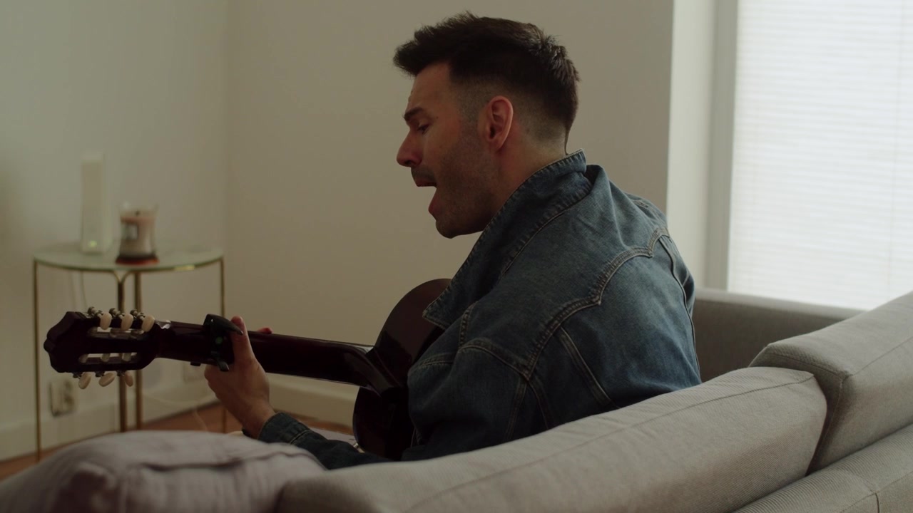 An authentic scene of a young man in a denim jacket playing a classic guitar while sitting on a sofa in a modern flat. Great for lifestyle and wellness content.