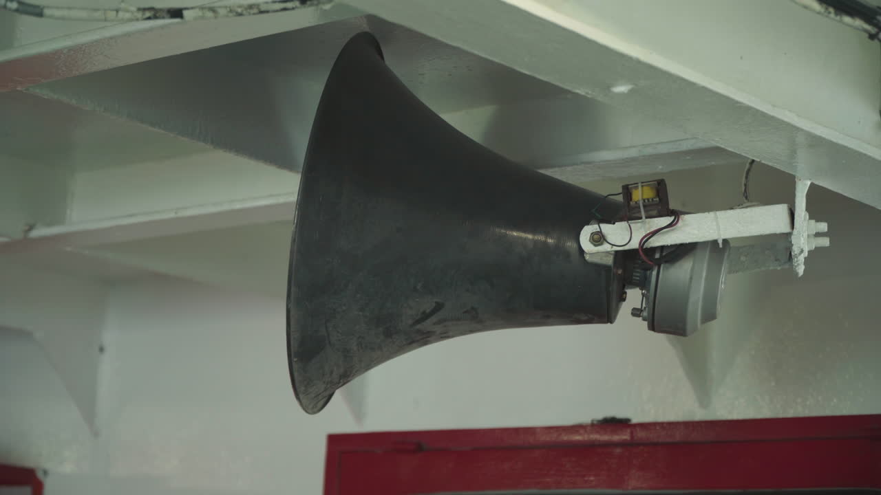 Tromba della nave montata sotto una trave del ponte, altoparlante conico nero fissato a una staffa bianca a soffitto all'interno della cabina, paratia rossa e supporto in acciaio visibili, primo piano che cattura corrosione, texture della vernice, bulloni