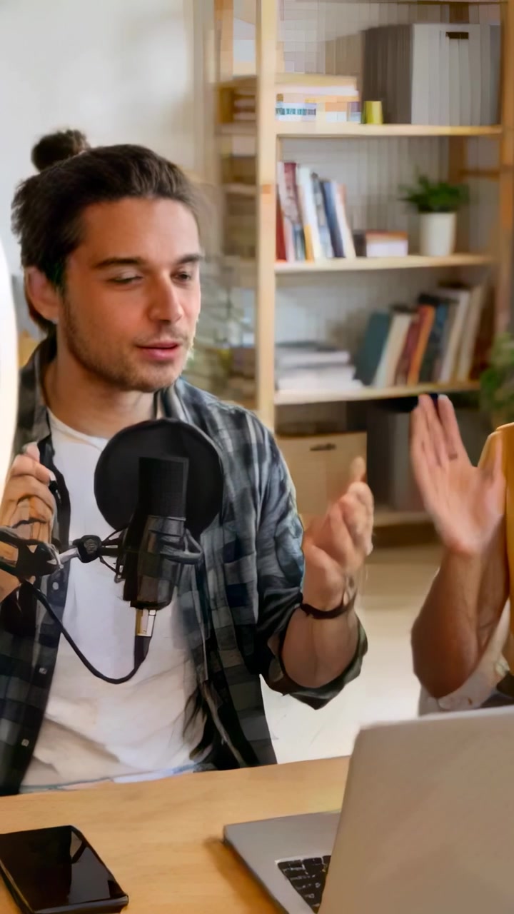 Video vertikal: Pria berkaus kotak-kotak menekan rekam, memperbaiki mikrofon dengan pop filter di meja untuk merekam sambil memberi isyarat dengan rekan pembawa acara.