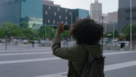 portrait of beautiful young african american woman taking photo of city building using smartphone enjoying urban sightseeing travel wearing backpack