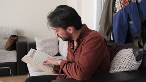 young muslim man sitting on the couch while reading holy book quran, people follow the rules of their religion