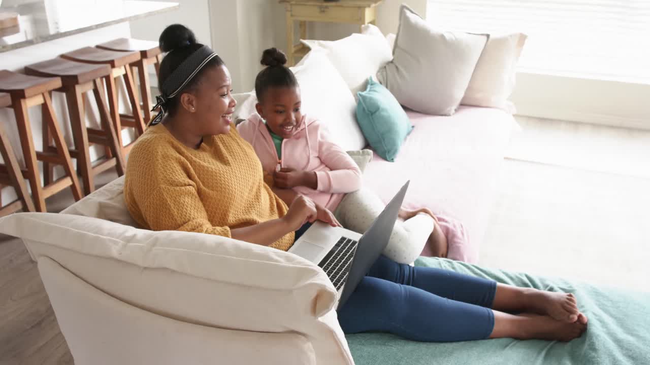 Happy african american mother and daughter relaxing on sofa using laptop, in slow motion