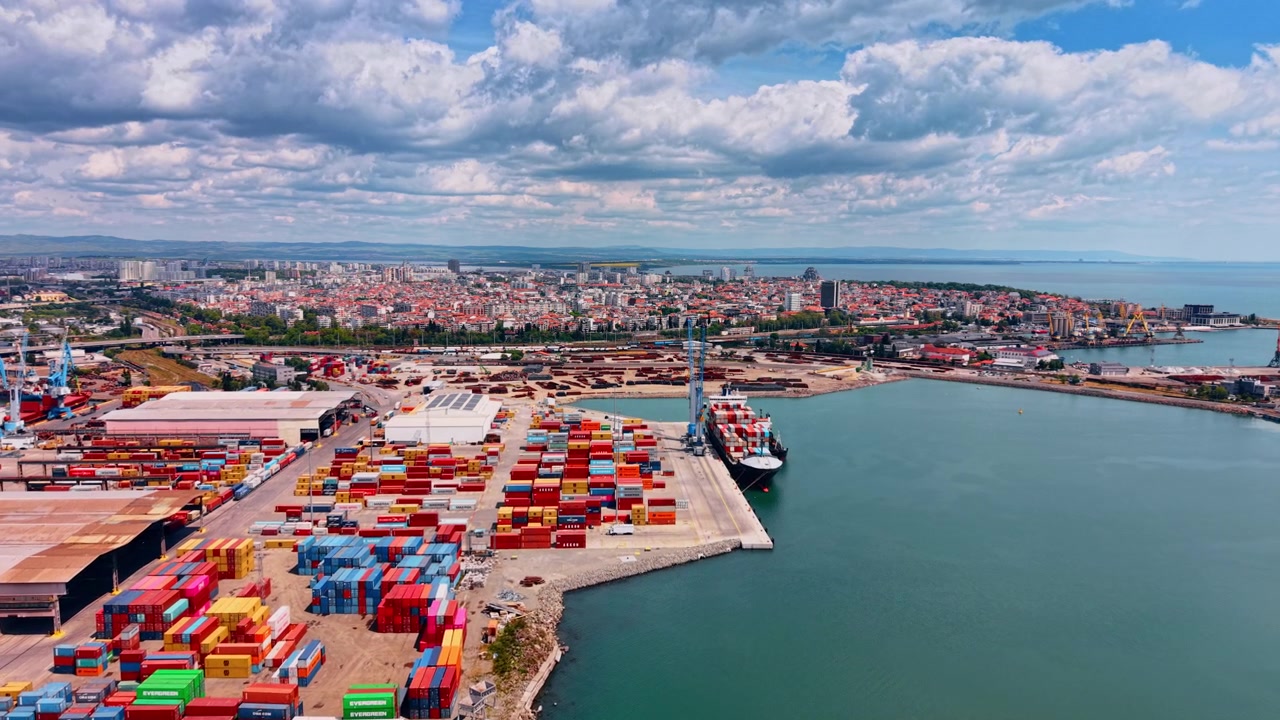 Burgas, Bulgaria, 1 May 2026: Aerial View of Container Terminal and Coastal City. Drone view of container yard, docked cargo ship, cranes, coastal city and sea under cloudy sky.