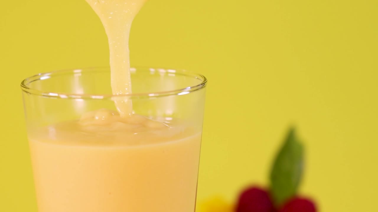 Un frullato di mango giallo denso e cremoso si versa in un bicchiere su uno sfondo giallo vibrante.