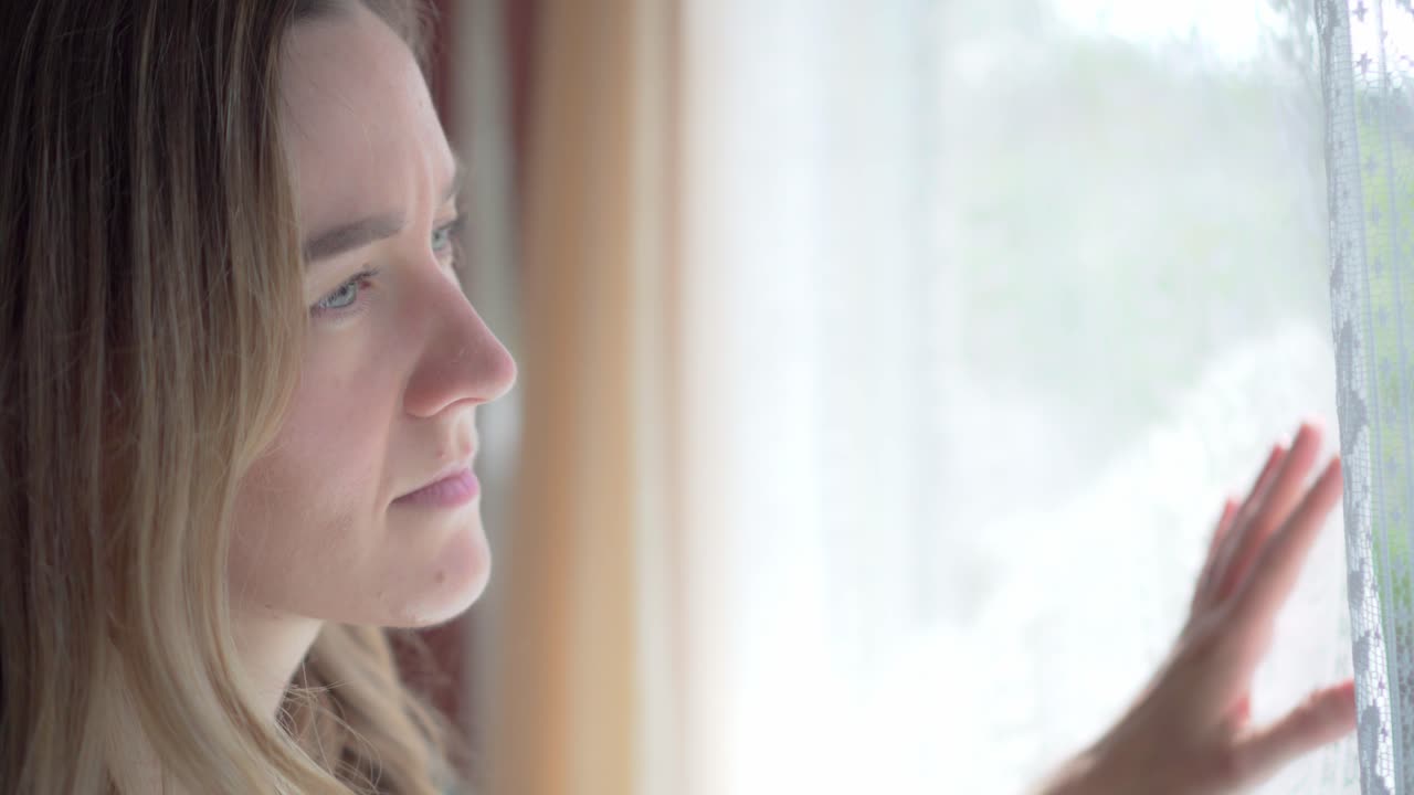 Sad girl inside house looks out through window with white lace curtains, close up