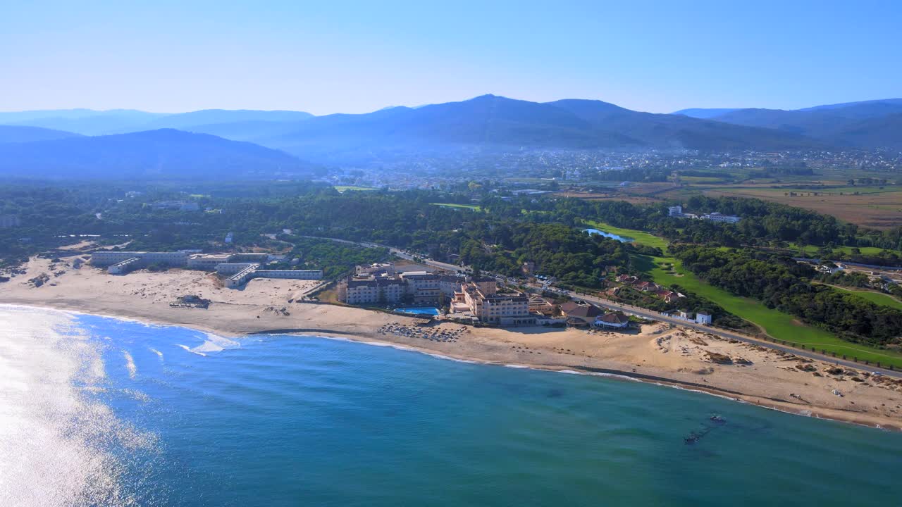 The scene is taken from a helicopter's point of view with the ocean in focus and mountains on the horizon. A beach bustles below while the water appears calm.