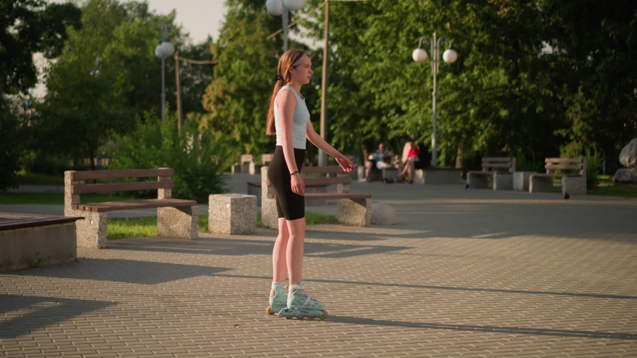 una mujer joven patinando lentamente al aire libre en un camino de pavimento entrelazado, rodeada de bancos de parque, vegetación y personas borrosas sentadas en el fondo