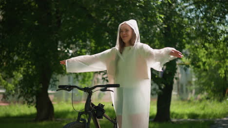 young woman in translucent raincoat stands by bicycle with arms joyfully outstretched, swinging her hands as if embracing freedom, sunlight filters through lush greenery