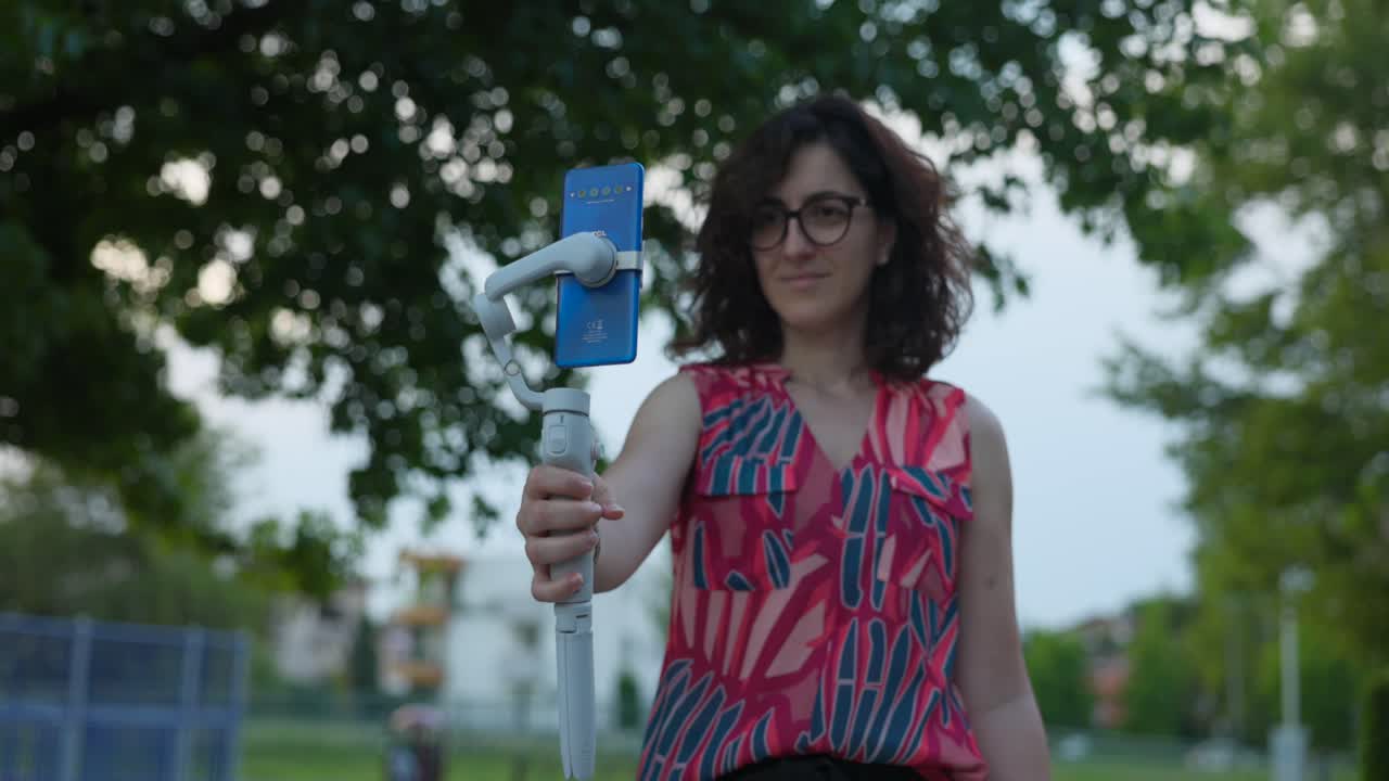 mujer con gafas sostiene osmo estabilizador gimbal móvil con teléfono celular montado al aire libre en el parque