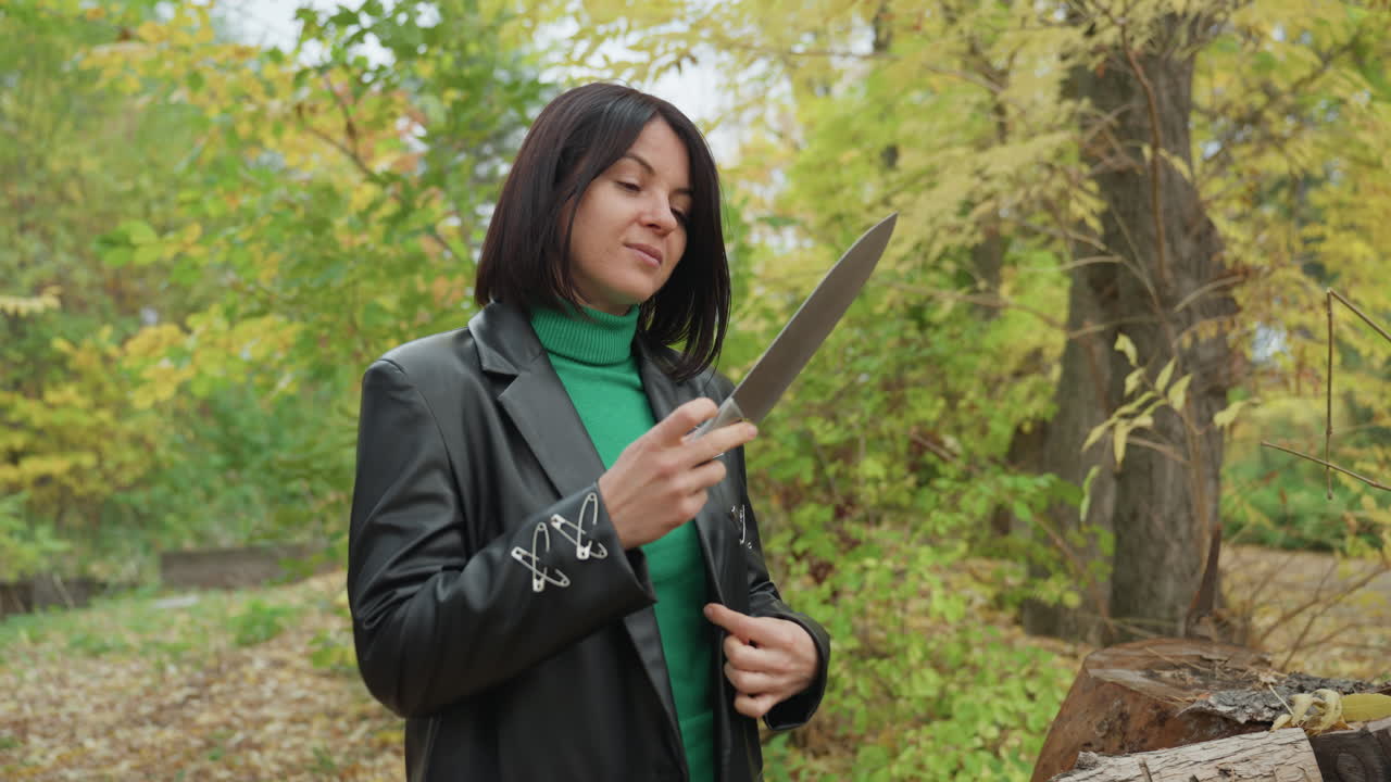 Frau mit Lederjacke inspiziert draußen zwischen Herbstbäumen ein Messer neben gestapelten Baumstämmen und einer Tasche, klappt die Klinge auf und studiert die Schneide mit konzentriertem Blick, düstere, einsame Atmosphäre vereint robusten Stil