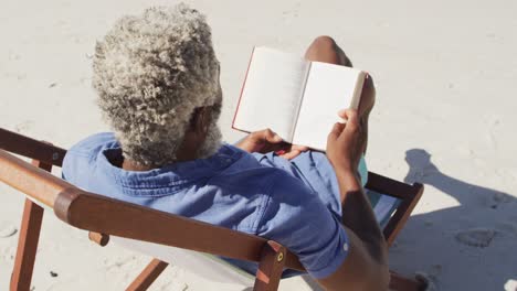hombre afroamericano mayor leyendo y acostado en una cama de sol en una playa soleada