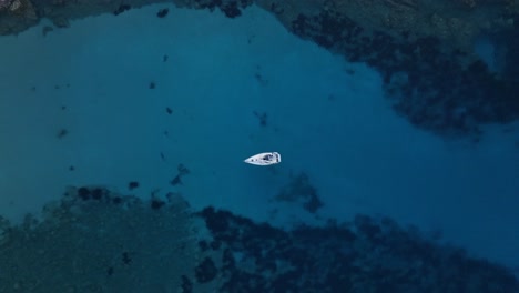 Drone-top-down-rising-above-sailboat-in-deep-blue-ocean-water,-rocky-limestone-cliffs-on-edge