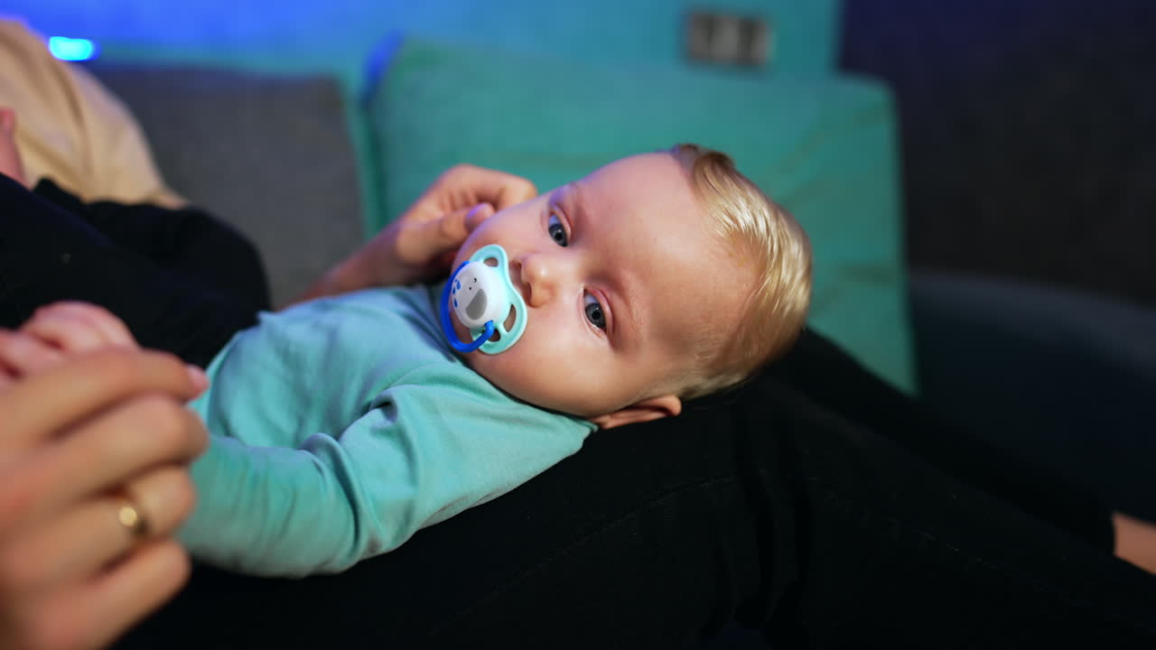 Lovely child with fair hair lies on mom's laps. Mother takes a pacifier out of kid's mouth and waves it in front of child's face. Close up.