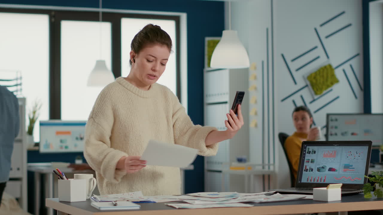Kvinde bruger smartphone i videokonference, taler om salg og holder papir med diagrammer