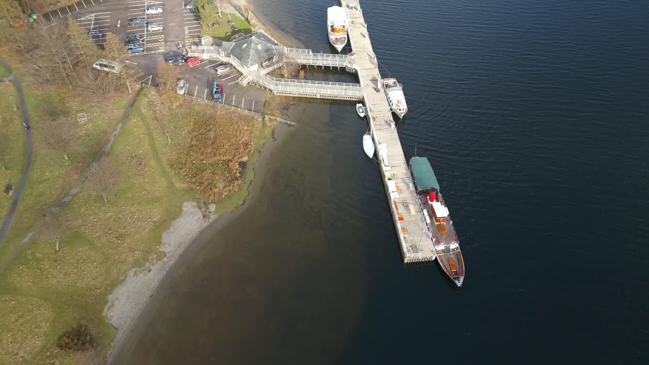 Retreating drone shot of a jetty where different kinds of boats are docked by the shore of the Ullswater Lake in Cumbria, Englan in United Kingdom