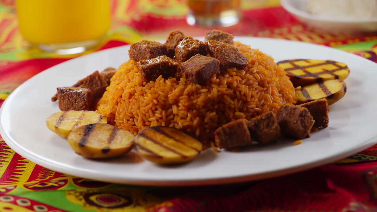 Tampilan dekat sepiring nasi yang disajikan dengan potongan daging kecoklatan dan irisan pisang raja panggang di atas taplak meja bermotif warna-warni, dengan minuman buram dan hidangan lain di latar belakang di bawah cahaya dalam ruangan yang hangat.