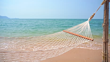 empty hammock on a sandy beach near a calm ocean with waves slowly reaching the shore during hot sunny day