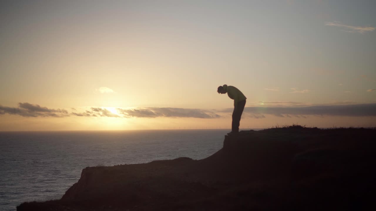 Silhouetted Man Sits on Cliff Overlooking Sunset at Sea