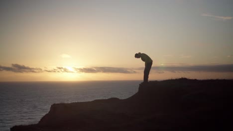 silhouetted man sits on cliff overlooking sunset at sea