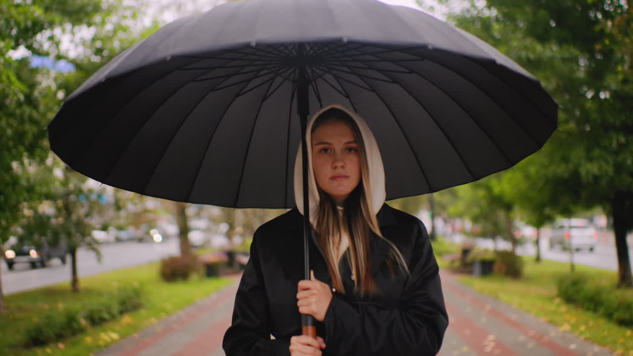 Woman in long black trench coat holding umbrella standing on city walkway during rainy autumn day, trees and cars in background, overcast sky, urban outdoor scene with calm mood and seasonal atmosphere