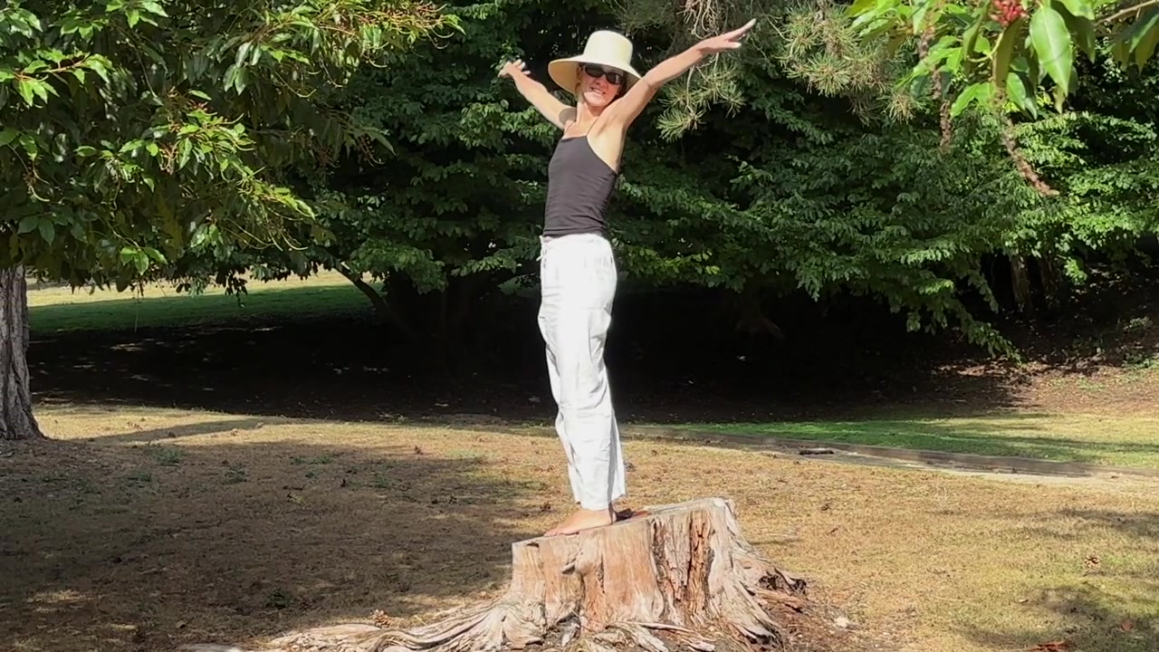 Donna con cappello da sole si alza e posa su un ceppo d'albero in un parco all'aperto. Inquadratura ampia