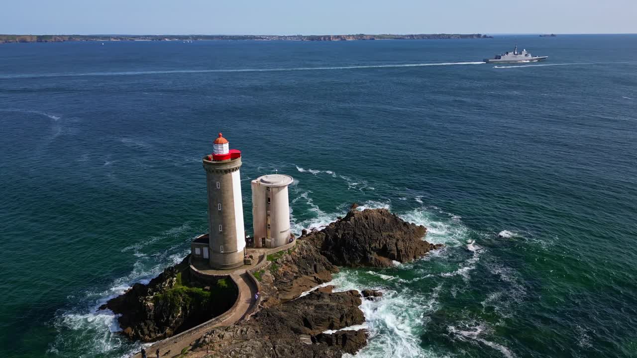 Drone orbiting around Petit Minou lighthouse in Brittany, revealing the rocky shore and path as a military ship crosses the open ocean in the background - Plouzané, France
