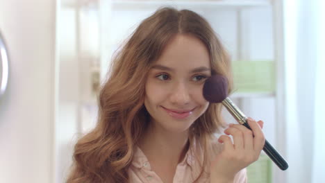 Young-woman-using-cosmetic-brush-for-applying-powder-on-face-at-bathroom-mirror