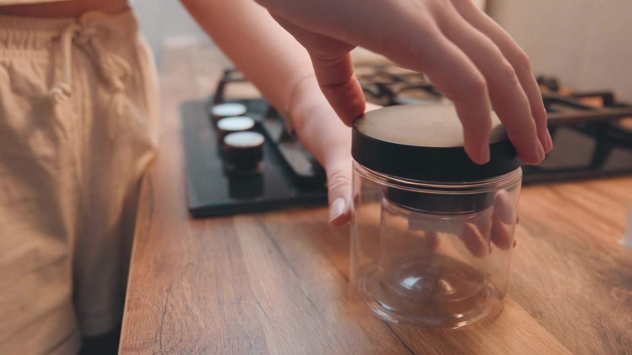 Jar sealing process. Individual gently twists black lid onto glass jar during meal preparation. Focused individual carefully secures black lid onto glass jar in welcoming kitchen setting