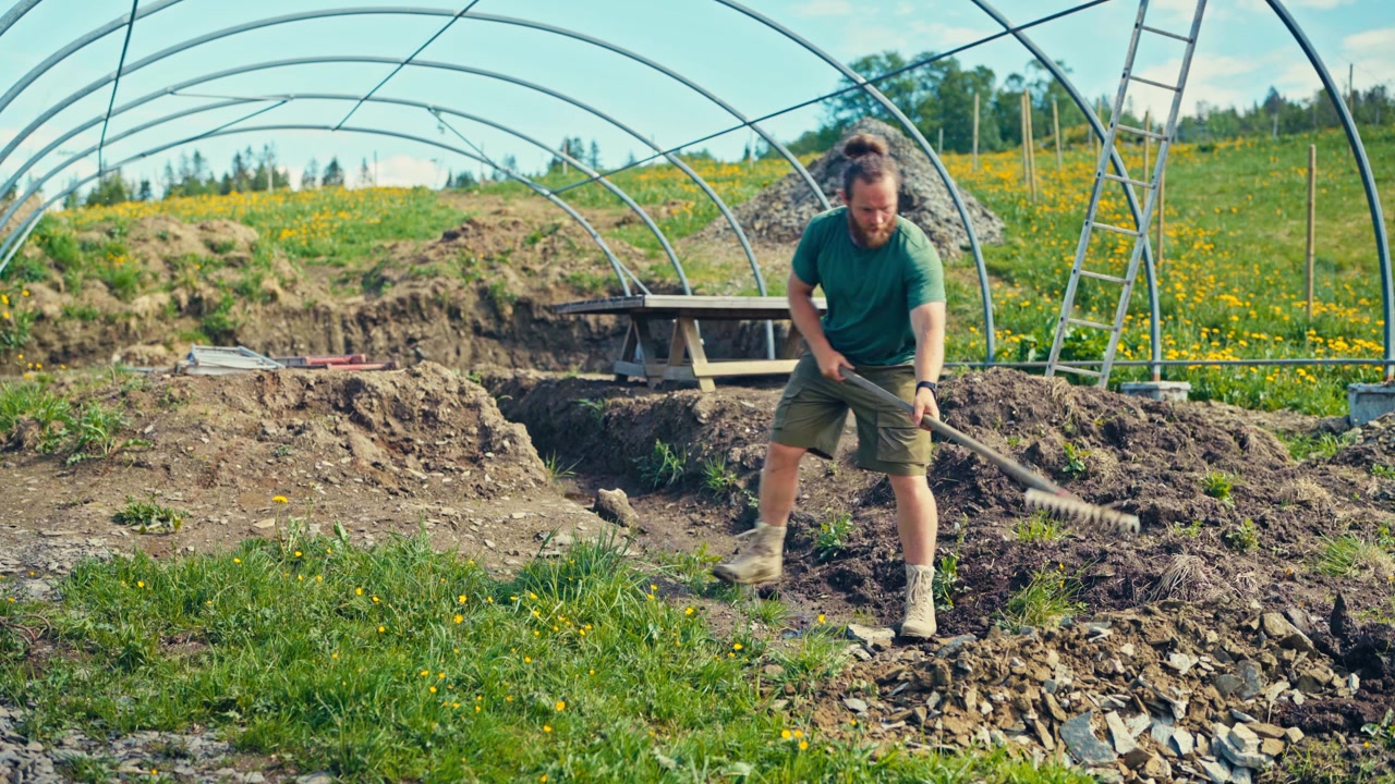 Man installing a greenhouse or polytunnel structure on a farm, showing agricultural work, construction materials, and the rural farming environment