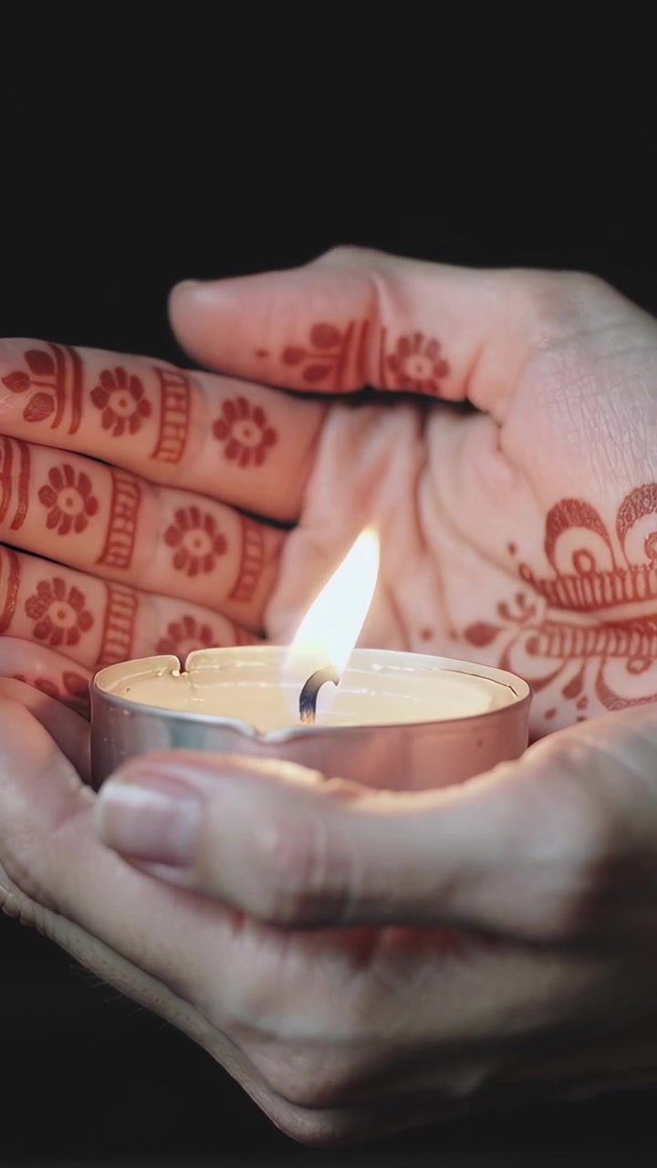 Vertical video: Reacting to flicker, mehndi hands steadying tealight in metal cup against black