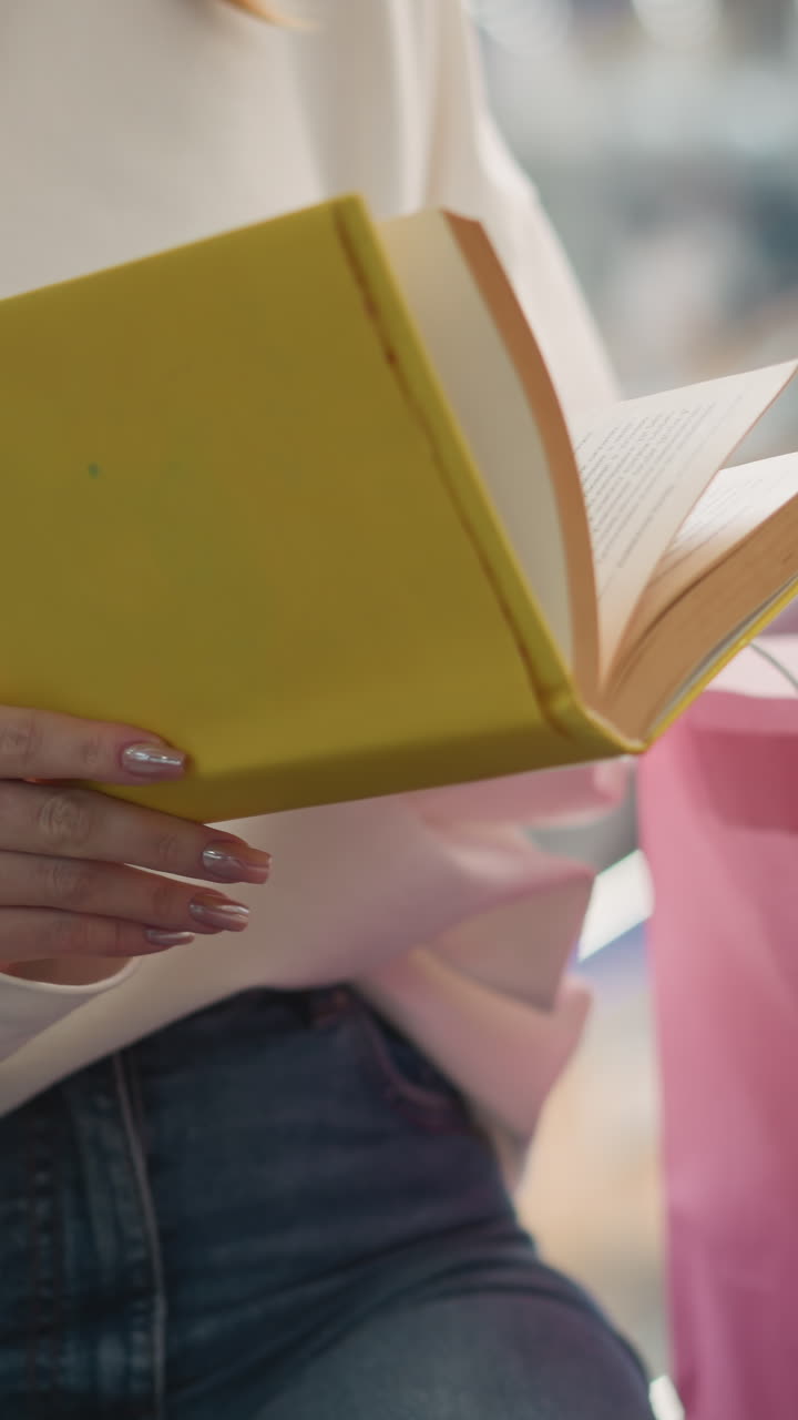 primer plano de una mujer sentada en el interior, hojeando un libro cubierto de amarillo con bolsas de compras de colores a su lado, capturando un momento casual y relajado en un entorno de centro comercial moderno y brillante